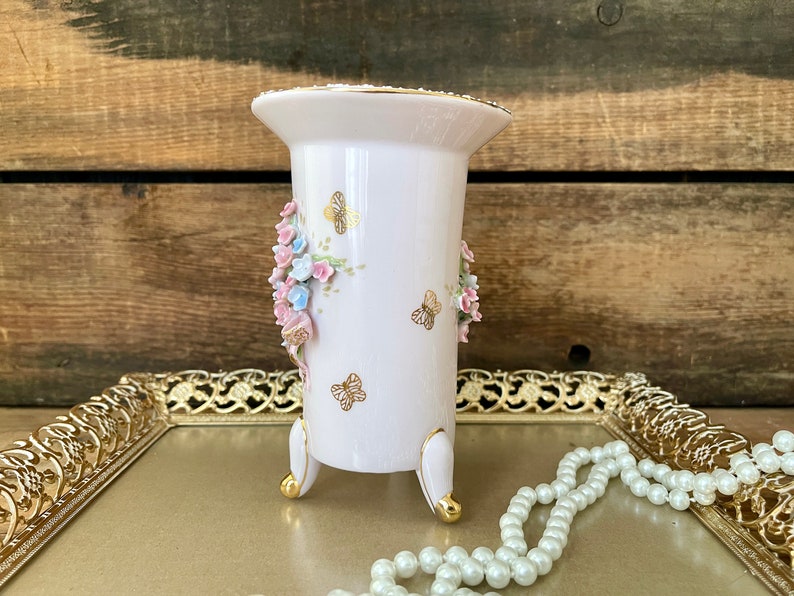 Peut inclure: Un vase en porcelaine blanche avec des accents dor&eacute;s et un motif floral compos&eacute; de fleurs roses et bleues et de papillons. Le vase a trois pieds dor&eacute;s et est pos&eacute; sur un plateau dor&eacute; avec un collier de perles.