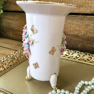 Peut inclure: Un vase en porcelaine blanche avec un motif floral et des accents dor&eacute;s. Le vase a trois pieds peints en or et est d&eacute;cor&eacute; de fleurs roses et bleues et de papillons.