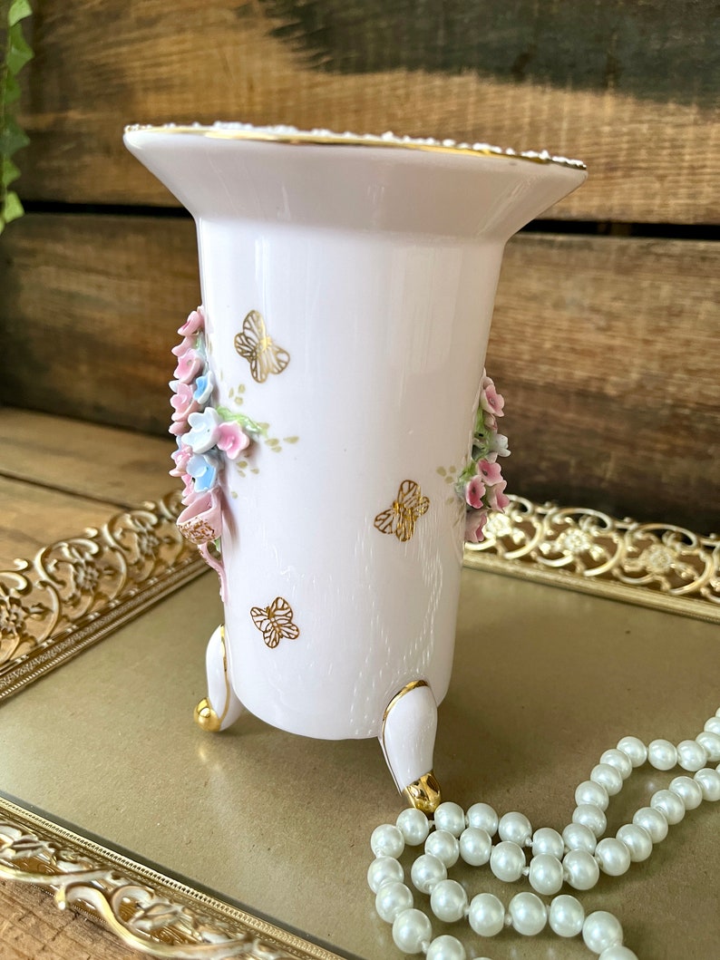 Peut inclure: Un vase en porcelaine blanche avec un motif floral et des accents dor&eacute;s. Le vase a trois pieds peints en or et est d&eacute;cor&eacute; de fleurs roses et bleues et de papillons.
