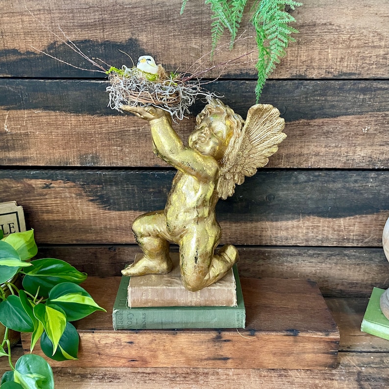 Puede incluir: Una estatua de querub&iacute;n de oro con alas arrodillada sobre una pila de libros. El querub&iacute;n sostiene un nido de p&aacute;jaro con un peque&ntilde;o p&aacute;jaro blanco dentro.