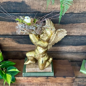 Puede incluir: Una estatua de querub&iacute;n dorada sosteniendo un nido de p&aacute;jaro con un p&aacute;jaro blanco dentro. La estatua est&aacute; sentada sobre una pila de libros.