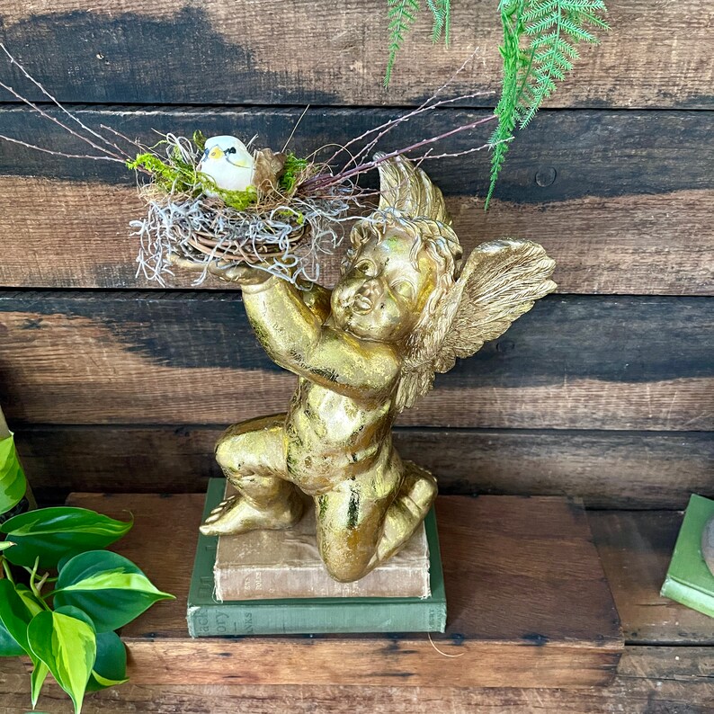 Puede incluir: Una estatua de querub&iacute;n dorada sosteniendo un nido de p&aacute;jaro con un p&aacute;jaro blanco dentro. La estatua est&aacute; sentada sobre una pila de libros.