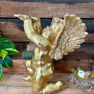 Puede incluir: Una estatua de querub&iacute;n dorada con las alas extendidas, arrodillada con la cabeza echada hacia atr&aacute;s. La estatua est&aacute; hecha de un material texturizado y tiene un acabado desgastado.