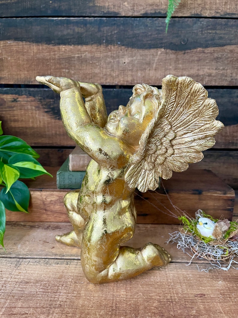 Puede incluir: Una estatua de querub&iacute;n dorada con las alas extendidas, arrodillada con la cabeza echada hacia atr&aacute;s. La estatua est&aacute; hecha de un material texturizado y tiene un acabado desgastado.