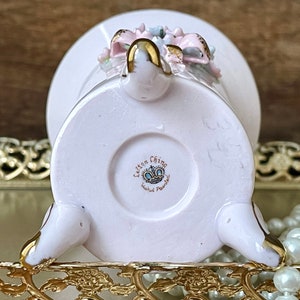 Peut inclure: Un vase en porcelaine rose avec un motif floral et des accents dor&eacute;s. Le vase a trois pieds et une base ronde. Le fond du vase porte le texte "Lefton China Hand Painted" avec une couronne.