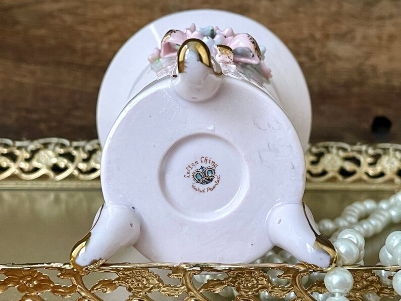 Peut inclure: Un vase en porcelaine rose avec un motif floral et des accents dor&eacute;s. Le vase a trois pieds et une base ronde. Le fond du vase porte le texte "Lefton China Hand Painted" avec une couronne.
