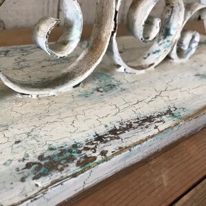 Op de afbeelding: Een wit houten plank met een verweerde afwerking en een decoratief metalen scrollwerk ontwerp. De plank heeft een lichtblauwe en gouden patina.