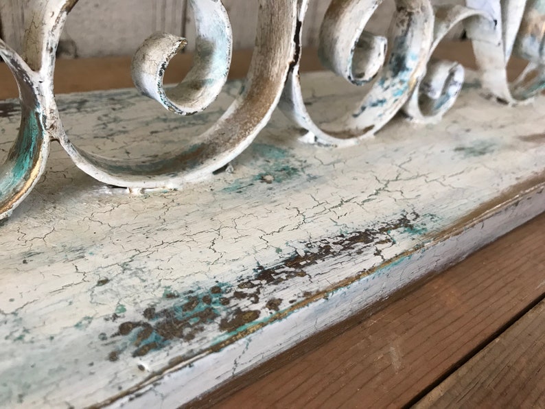 Op de afbeelding: Een wit houten plank met een verweerde afwerking en een decoratief metalen scrollwerk ontwerp. De plank heeft een lichtblauwe en gouden patina.