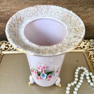 Peut inclure: Un vase en c&eacute;ramique rose avec un bord dor&eacute; et un motif floral. Le vase a trois pieds dor&eacute;s et est d&eacute;cor&eacute; de fleurs roses et bleues.
