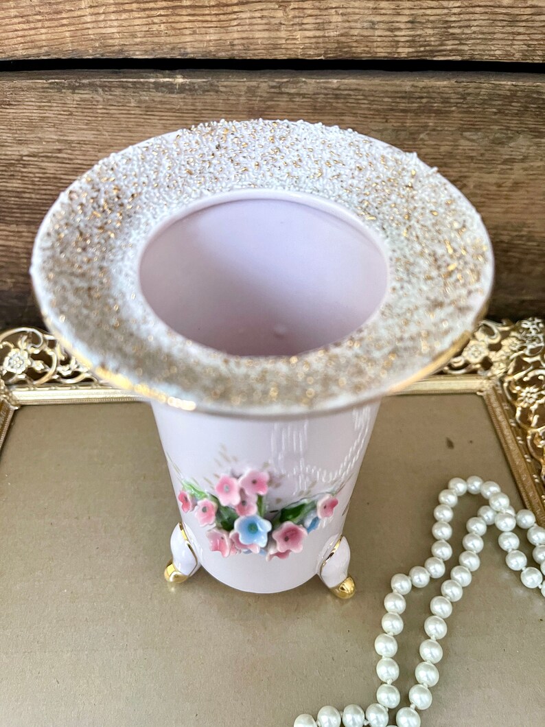 Peut inclure: Un vase en c&eacute;ramique rose avec un bord dor&eacute; et un motif floral. Le vase a trois pieds dor&eacute;s et est d&eacute;cor&eacute; de fleurs roses et bleues.