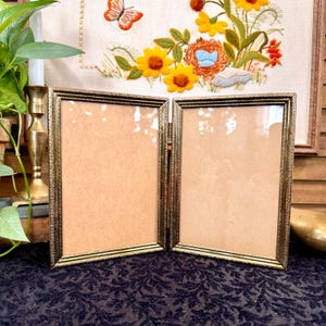 May include: A hinged double picture frame made of metallic gold-toned material with a textured, decorative border. The frame is designed to hold two vertical photographs and stands open on a flat surface, featuring clear glass panels and tan backings.