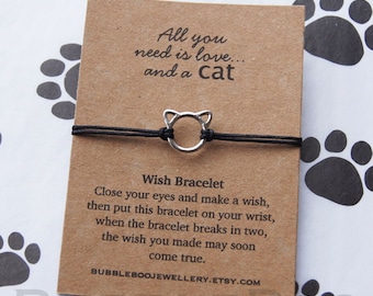 May include: A black string bracelet with a silver cat charm. The bracelet is on a card with the text "All you need is love... and a cat" and "Wish Bracelet". The card also includes instructions on how to make a wish with the bracelet.