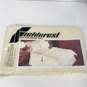 May include: A cream-coloured package of Fieldcrest full flat percale sheet set. The packaging features the Fieldcrest logo in black. The image on the packaging shows a cream-coloured bed set with decorative pillows and a bed skirt. The text "TROUSSEAU RIBBON" is also visible.