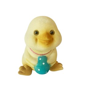 Peut inclure: Figurine de canard jaune avec un bec et des yeux brun clair. Le canard tient un objet bleu en forme d'œuf avec des pois blancs. Il a un collier rouge et des pieds bruns. La figurine est sur fond blanc.