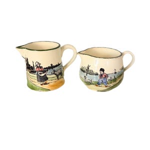 May include: Two cream-colored ceramic pitchers with hand-painted pastoral scenes. One pitcher depicts a woman with a basket and a sheep, the other a child with a dog. Both have green trim and curved handles.