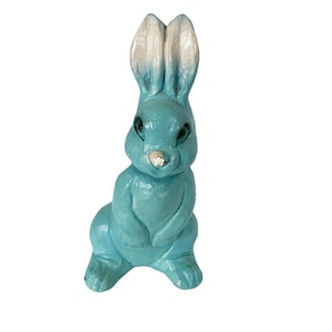 Peut inclure: Figurine de lapin bleu clair avec des oreilles à bout blanc. Le lapin est en position assise, les pattes jointes. La figurine a des yeux noirs et un petit nez doré.