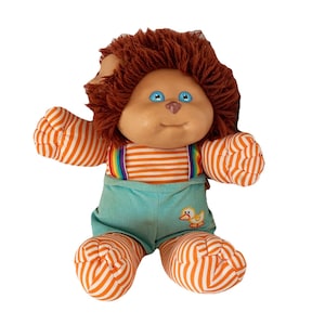 May include: A plush doll with brown yarn hair, blue eyes, and a small nose. The doll wears an orange and white striped long-sleeve shirt with rainbow suspenders and light teal corduroy overalls featuring a small embroidered yellow duck graphic.