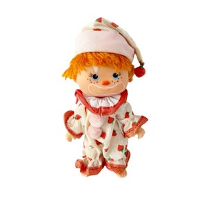 May include: A vintage clown doll with orange hair, blue eyes, and a painted smile. The doll wears a white outfit with red strawberry patterns and trim, a ruffled collar, and a matching hat. The hat has a red pom-pom.