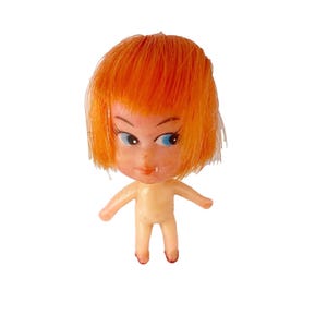 May include: A small plastic doll with orange hair and blue eyes. The doll is wearing no clothes and has a simple, childlike face.