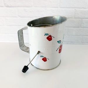 May include: White vintage sifter with a metal handle and rim. The sifter has red apple illustrations and measurement markings. The sifter has a crank with a black handle. A classic kitchen tool for baking.
