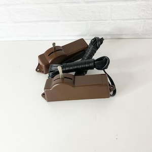 Puede incluir: Dos controladores de velocidad de motor eléctrico marrones vintage con cables negros. Cada controlador tiene una palanca y una base rectangular. Los cables están enrollados y conectados a los controladores. El fondo es blanco con una pared de ladrillos blancos.