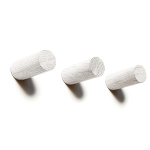 May include: Three white wooden hooks mounted on a white wall. The hooks are cylindrical in shape and are arranged in a row.