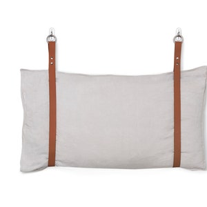 Könnte beinhalten: Ein hellgraues Leinenkissen mit zwei braunen Lederriemen, die an Metallhaken befestigt sind. Das Kissen hängt an den Haken.