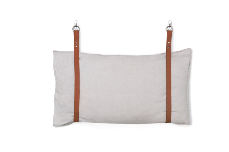 May include: A light grey linen pillow with two brown leather straps attached to metal hooks. The pillow is hanging from the hooks.