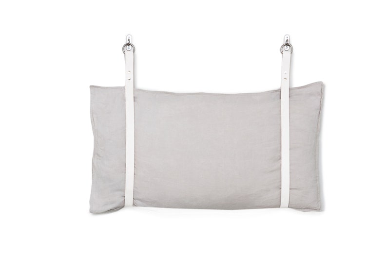 May include: A light grey linen pillow with white leather straps and silver metal hooks. The pillow is hanging from the straps.