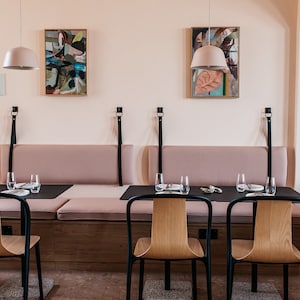 May include: A dining area with a long, cushioned bench and two black chairs. The bench has black leather straps and the chairs have wooden seats. There are two abstract paintings on the wall and two pendant lights hanging above the table. The walls are painted a pale pink.