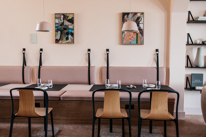 May include: A dining area with a long, cushioned bench and two black chairs. The bench has black leather straps and the chairs have wooden seats. There are two abstract paintings on the wall and two pendant lights hanging above the table. The walls are painted a pale pink.