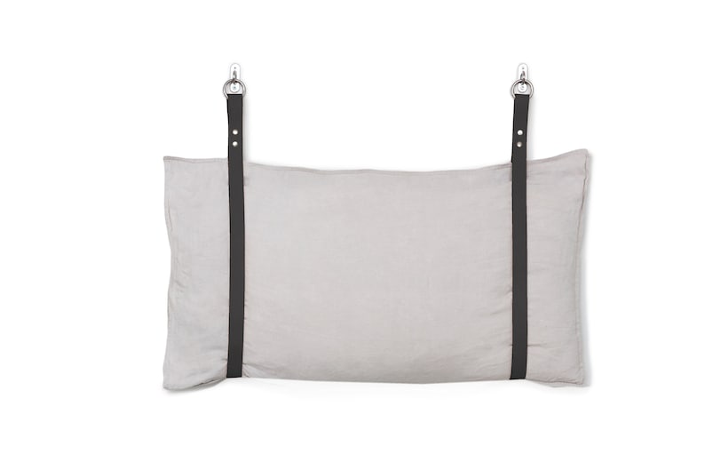 May include: A light grey linen pillow with two black straps and silver hooks attached to the top.