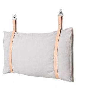 May include: A light grey linen pillow with two tan leather straps attached to metal hooks. The pillow is hanging on a white wall.