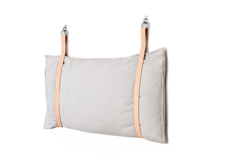 May include: A light grey linen pillow with two tan leather straps attached to metal hooks. The pillow is hanging on a white wall.