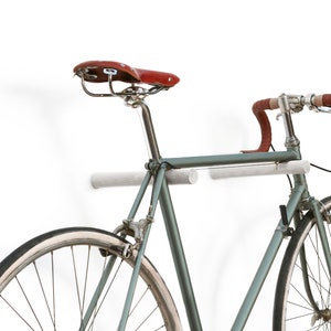 Puede incluir: Una bicicleta gris con manillar y sillín de cuero marrón está montada en una pared blanca utilizando un par de ganchos cilíndricos blancos.