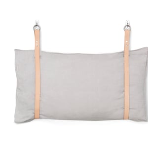 May include: A grey linen pillow with two leather straps attached to metal hooks. The pillow is hanging on a white wall.