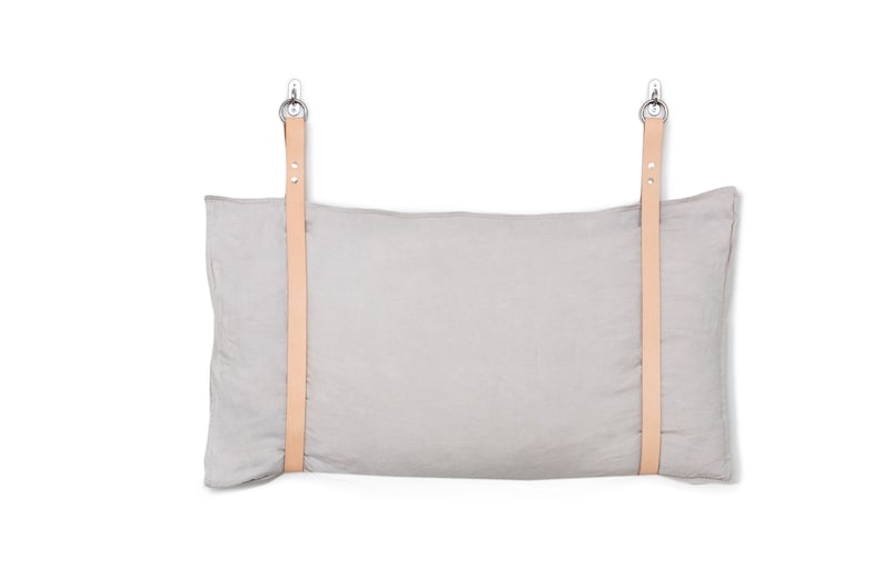 May include: A grey linen pillow with two leather straps attached to metal hooks. The pillow is hanging on a white wall.