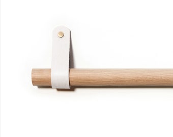 Puede incluir: Una barra de madera con una correa de cuero blanco y un botón de latón. La barra está montada en una pared blanca.
