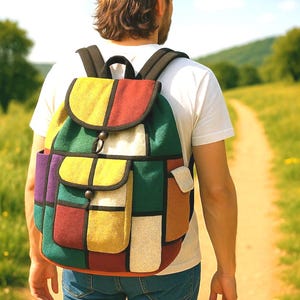 May include: A vibrant patchwork backpack featuring panels of red, yellow, green, purple, and beige. The bag has black straps and a wooden toggle. The person is wearing a white t-shirt, blue jeans, and a beige baseball cap. The person is walking on a path.
