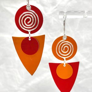 May include: Geometric dangle earrings featuring a red circle with a silver spiral, connected to an orange triangle and a red circle. The earrings have silver hooks and a modern, abstract design.