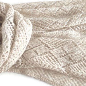 May include: A light beige, sheer knitted scarf featuring an intricate openwork diamond pattern. The fabric has a delicate, airy texture with a soft, fuzzy appearance, showcasing a repeating geometric lattice design throughout the entire length.