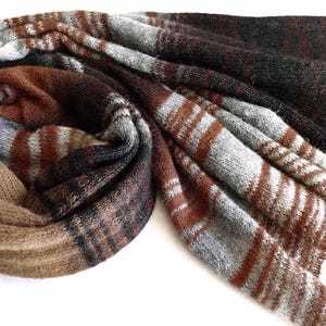 Könnte beinhalten: Ein langer, rechteckiger Strickschal mit einem mehrfarbigen Streifenmuster in den Farben Dunkelbraun, Hellbraun, Grau und Weiß. Der Stoff hat eine weiche, strukturierte Optik mit horizontalen Streifen in unterschiedlichen Breiten und Farben.