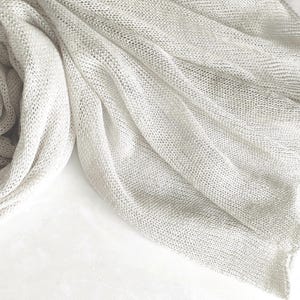 May include: A lightweight, off-white scarf featuring a loose, open-knit construction. The fabric displays a semi-sheer, airy texture with soft, flowing folds that create natural drapes across the surface, showcasing a consistent, fine-gauge weave.