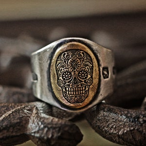 May include: A silver ring with a gold-toned oval inlay featuring a detailed sugar skull design.