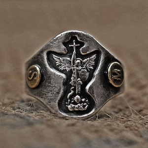 Op de afbeelding: Een zilveren ring met een gedetailleerd ontwerp van een engel die een zwaard vasthoudt en op een kruis staat. De ring heeft twee gouden accenten aan de zijkanten, een met de letter "S" en de andere met de letter "W".