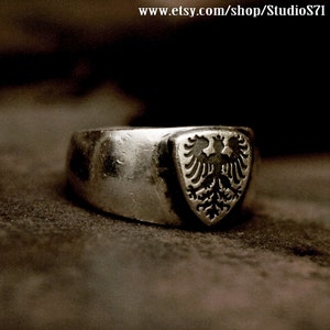 May include: A silver ring with a shield-shaped design featuring a black double-headed eagle.