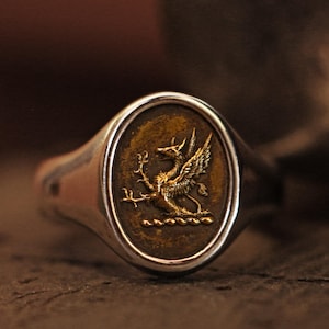 May include: A silver ring with an oval-shaped setting featuring a gold-colored griffin crest.