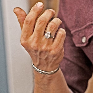 Op de afbeelding: Een zilveren zegelring met een bloemmotief wordt aan een hand gedragen, samen met een zilveren armband. De voorkant van de ring heeft een gedetailleerd, reliëfpatroon. De armband is een eenvoudige, open manchet. De hand is tegen een bordeauxrode achtergrond.