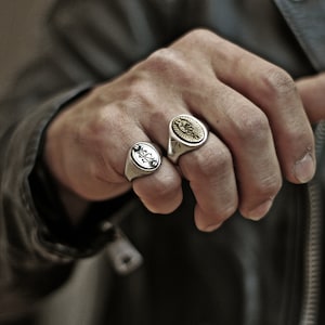 May include: Two silver oval rings with engraved designs. One ring features a sun with a face and the other ring has a floral design.