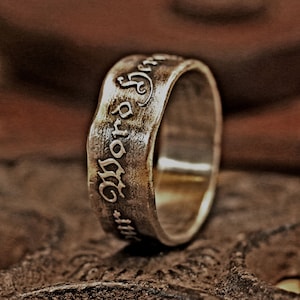 May include: A silver ring with an engraved inscription. The ring has a textured surface and a slightly irregular shape. The inscription appears to be in an old English font. The ring is displayed on a brown surface.